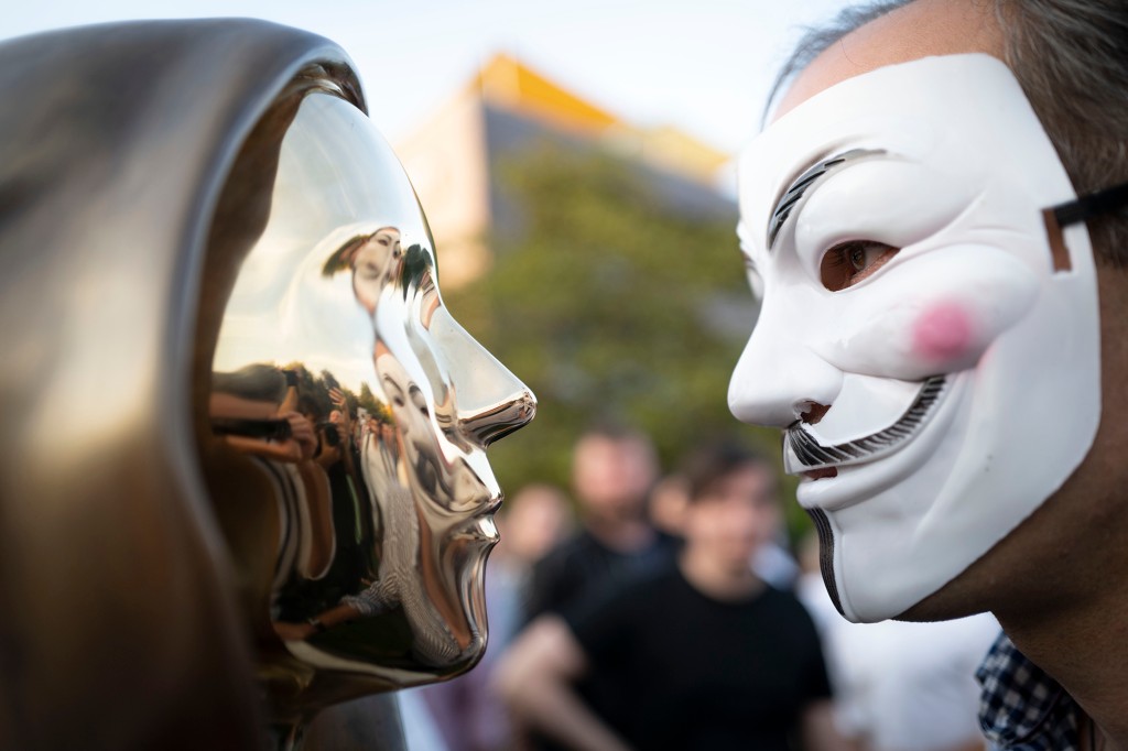 Un hombre con una máscara de Guy Fawkes mira una estatua del misterioso desarrollador de la moneda digital Bitcoin en Budapest.