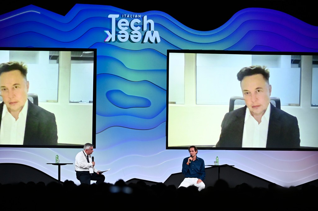 Elon Musk visto en dos pantallas grandes debajo de las palabras TechWeek mientras dos personas se sientan en el escenario debajo de las pantallas.