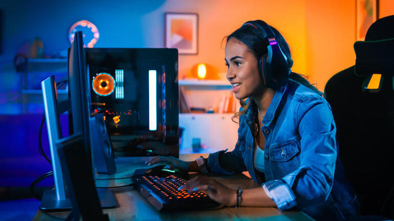 Bonita y emocionada Black Gamer Girl con auriculares está jugando un videojuego de disparos en primera persona en línea en su computadora.  La habitación y la PC tienen luces LED de neón de colores.  Noche acogedora en casa.
