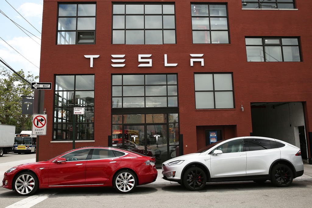 Un vehículo Tesla rojo y un vehículo Tesla blanco estacionado en la calle fuera de un edificio de ladrillo con TESLA escrito en el exterior