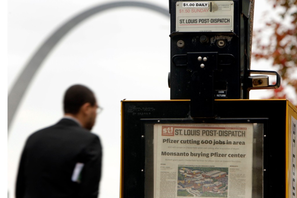 Un hombre pasa junto a una caja de periódicos que contiene el periódico St. Louis Post Dispatch.  Al fondo se ve una parte del arco St. Louis Gateway.