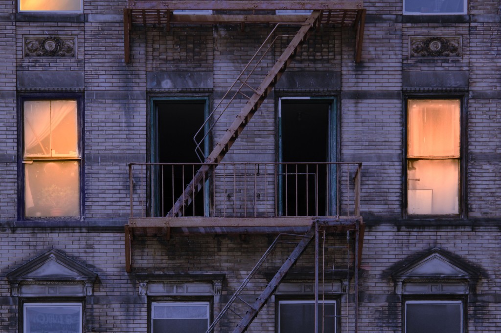 Vista nocturna de la fachada de ladrillo erosionado de un edificio residencial sin ascensor en la ciudad de Nueva York con una escalera de incendios oxidada y ventanas de guillotina viejas