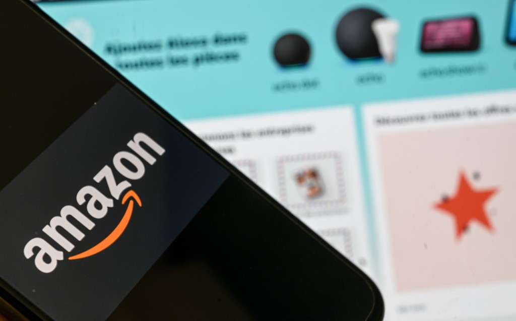 una persona posa con un teléfono inteligente que muestra un logotipo de Amazon, frente a una pantalla de computadora que muestra una página de inicio de Amazon